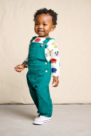 Young child wearing green overalls and patterned long sleeve shirt standing against a textured backdrop
