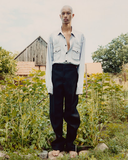 Young adult poses barefoot in a light blue button-down and black pants in a verdant rural setting with a weathered barn backdrop.