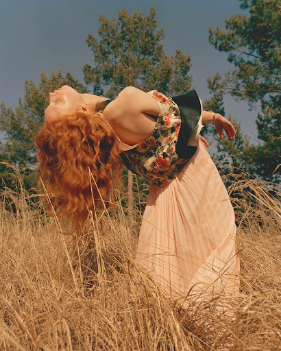 A woman with red hair leans back in a field of tall grass, wearing a floral top and light pink skirt against a backdrop of trees.