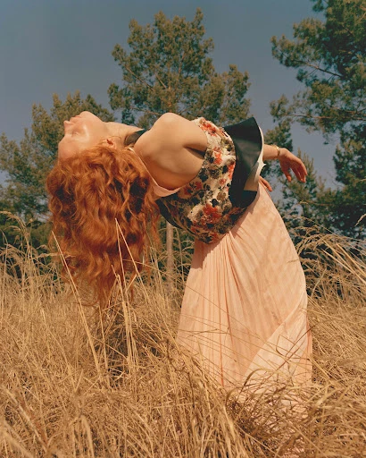 A woman with red hair leans back in a field of tall grass, wearing a floral top and light pink skirt against a backdrop of trees.