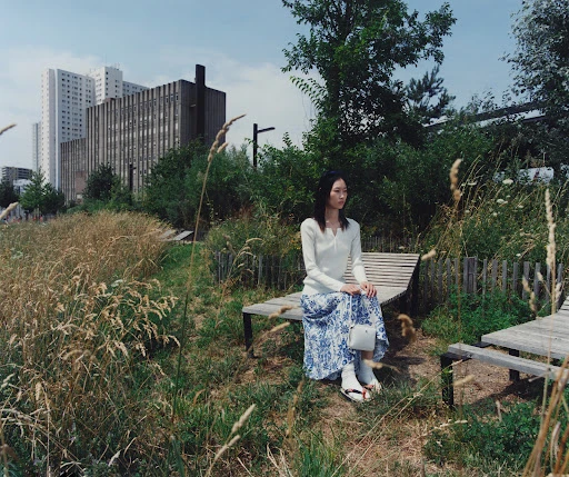 Asian woman seated on a wooden lounger in a park wearing a white top and blue patterned skirt