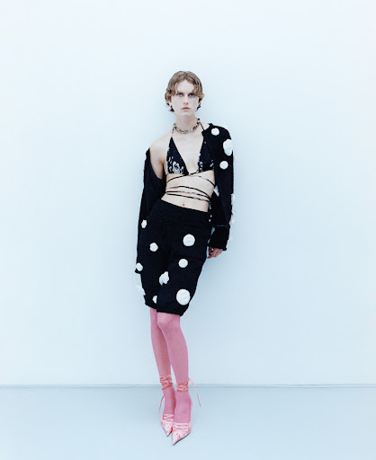 Full-length shot of a stylish young adult female in a black polka-dot outfit and pink stockings, posed against a light blue background.