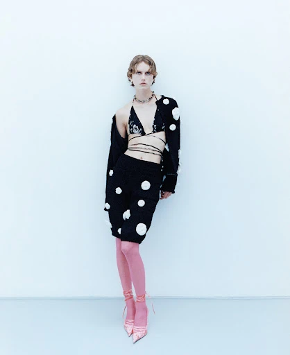 Full-length shot of a stylish young adult female in a black polka-dot outfit and pink stockings, posed against a light blue background.
