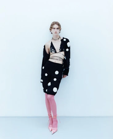 Full-length shot of a stylish young adult female in a black polka-dot outfit and pink stockings, posed against a light blue background.