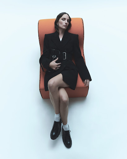 A young adult woman lounges in an orange chair wearing a black belted blazer dress with black shoes and a black purse against a white background.