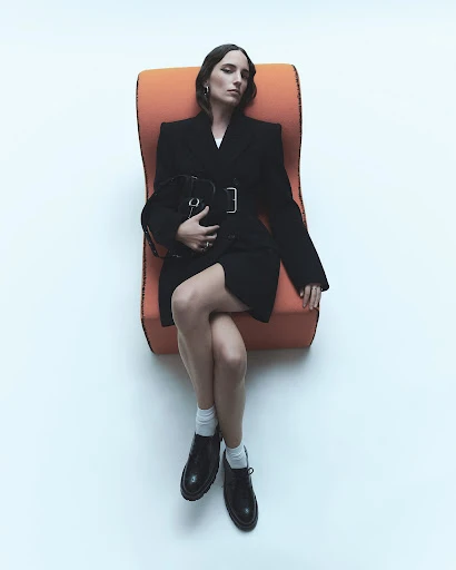 A young adult woman lounges in an orange chair wearing a black belted blazer dress with black shoes and a black purse against a white background.