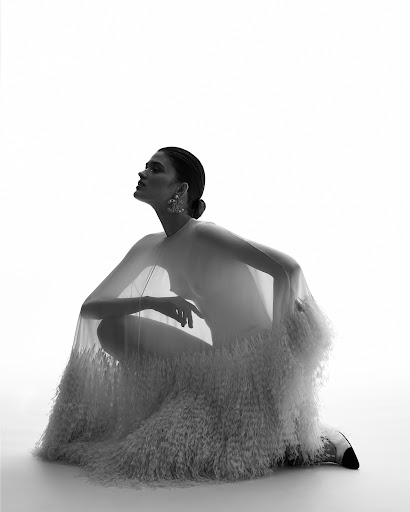 Monochrome fashion portrait of a woman in a sheer textured gown crouching in a bright white studio environment, looking upwards in soft light.