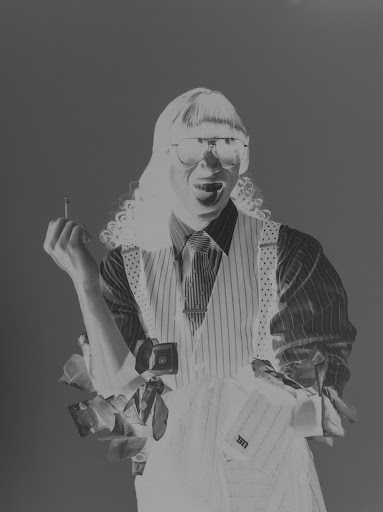 Person in glasses and vest holds a cigarette and collared shirts against a blurred background in a black and white photograph.