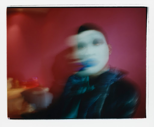 Abstract polaroid of a person with makeup in a dark jacket against a red wall, creating a vintage and artistic effect.