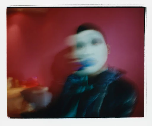 Abstract polaroid of a person with makeup in a dark jacket against a red wall, creating a vintage and artistic effect.