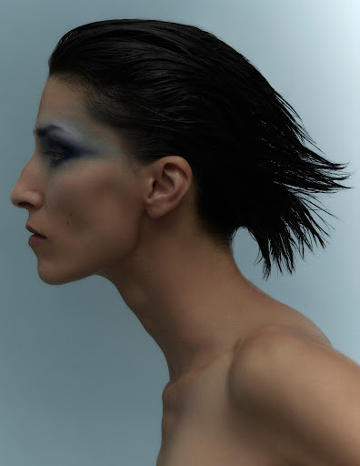 Profile portrait of a woman with slicked-back black hair and blue eyeshadow against a pale blue background in a beauty shot.