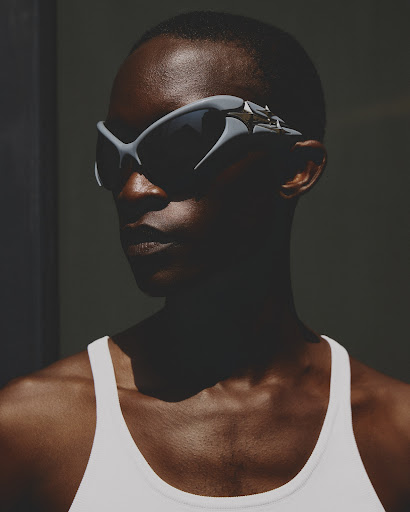 Close-up of a dark-skinned woman wearing angular gray sunglasses and a white top against a dark gray background in studio lighting.