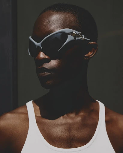 Close-up of a dark-skinned woman wearing angular gray sunglasses and a white top against a dark gray background in studio lighting.