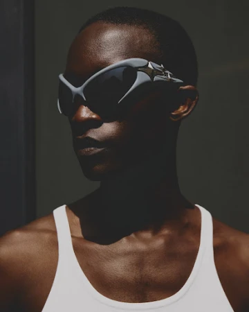 Close-up of a dark-skinned woman wearing angular gray sunglasses and a white top against a dark gray background in studio lighting.
