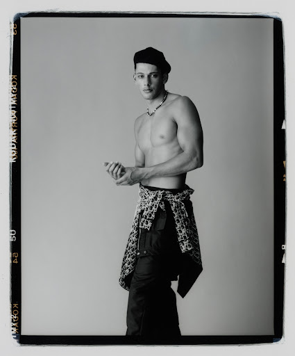 Black and white portrait of a shirtless man wearing a beret and patterned pants tied around his waist against a grey background.