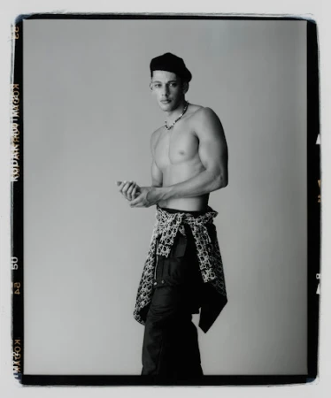 Black and white portrait of a shirtless man wearing a beret and patterned pants tied around his waist against a grey background.