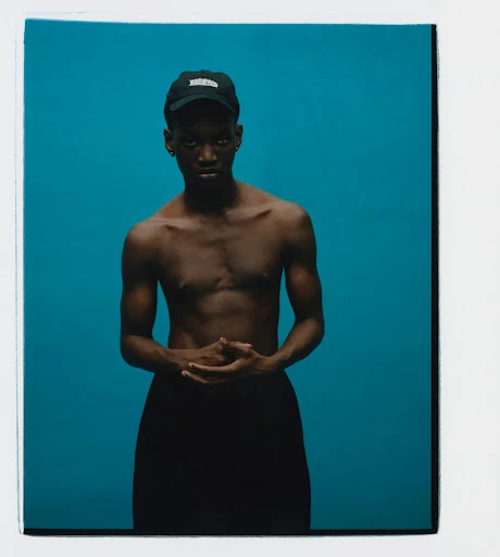 Studio portrait of a bare-chested young Black man in a black cap, set against a vibrant teal backdrop. He wears black trousers.
