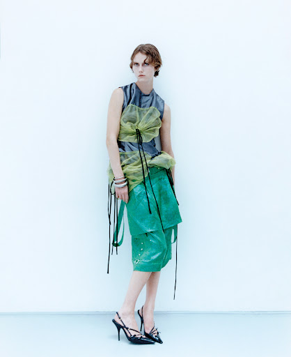 A slender adult fashion model poses indoors against a white wall wearing a sleeveless gray top, green culottes, and strappy high heels.