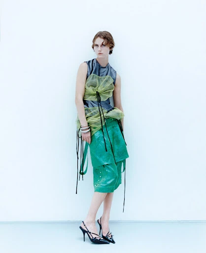 A slender adult fashion model poses indoors against a white wall wearing a sleeveless gray top, green culottes, and strappy high heels.