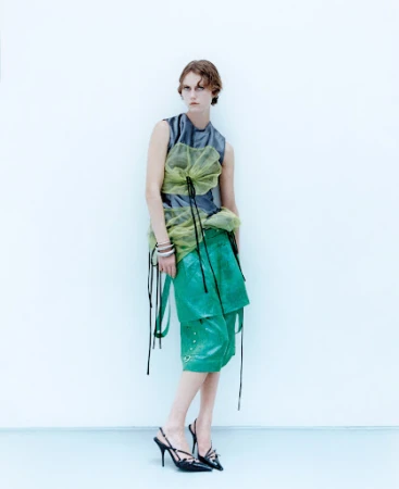 A slender adult fashion model poses indoors against a white wall wearing a sleeveless gray top, green culottes, and strappy high heels.