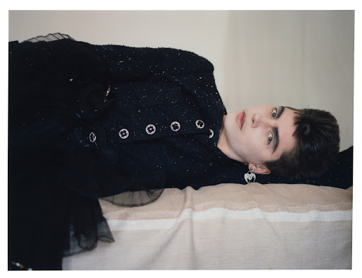 Fashion portrait of a young adult female model with dark eye makeup, a black jacket, and earrings, lying on a striped fabric surface.