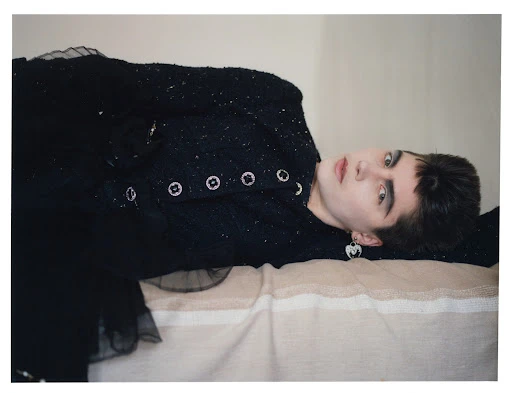 Fashion portrait of a young adult female model with dark eye makeup, a black jacket, and earrings, lying on a striped fabric surface.