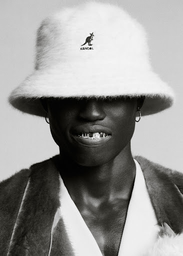 Monochrome portrait of a stylish adult male wearing a Kangol bucket hat, an unbuttoned robe, and grill jewelry with a neutral background.