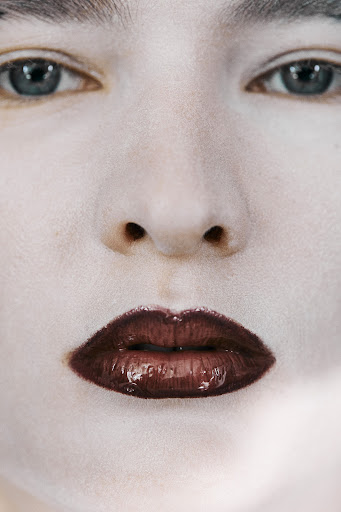 Close-up portrait of a person with fair skin, dark eyes, and bold, dark brown glossy lips, emphasizing makeup and facial features.