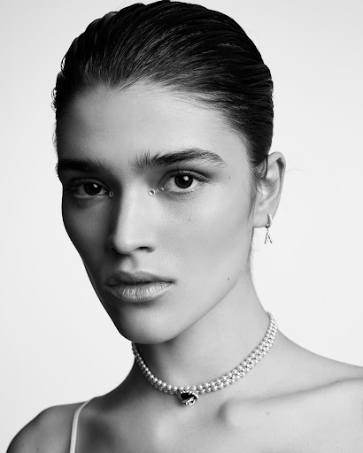 Monochrome portrait of a young woman with an under-eye piercing, wearing a pearl choker necklace, against a plain white backdrop.