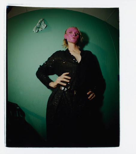 Woman with pink face mask poses indoors against teal wall in black sequin dress, hands on hips and wearing a belt, in a studio photo.