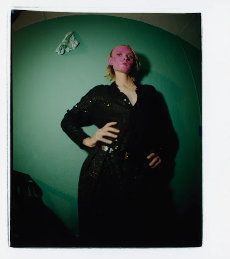 Woman with pink face mask poses indoors against teal wall in black sequin dress, hands on hips and wearing a belt, in a studio photo.