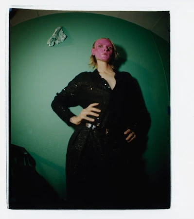 Woman with pink face mask poses indoors against teal wall in black sequin dress, hands on hips and wearing a belt, in a studio photo.