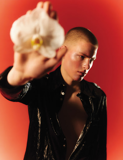 A young adult is wearing a black shirt and holding a white orchid flower against a red background in a studio lighting setting.