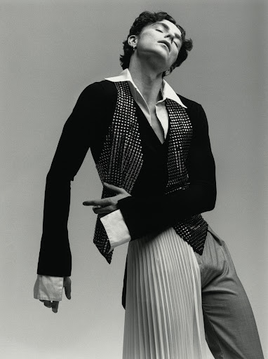 Fashion portrait of a curly-haired male model in a black jacket with a sequined vest, a white pleated skirt, and gray trousers against gray.