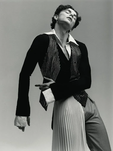 Fashion portrait of a curly-haired male model in a black jacket with a sequined vest, a white pleated skirt, and gray trousers against gray.