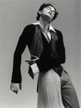 Fashion portrait of a curly-haired male model in a black jacket with a sequined vest, a white pleated skirt, and gray trousers against gray.