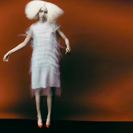 Model with voluminous white hair wearing a sheer layered white dress and orange heels against a rust backdrop