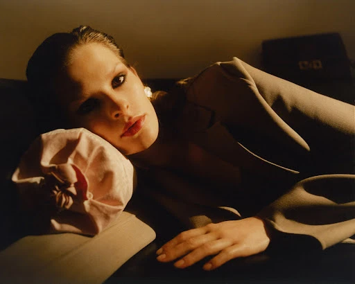Portrait of a woman lying on a tan couch wearing a suit jacket and earrings, with a neutral pillow behind her head.