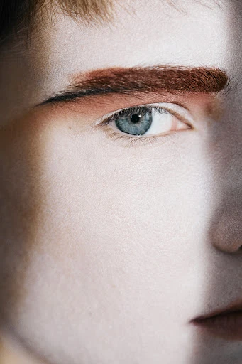 Close-up portrait of a young adult's face, featuring a light skin tone, striking blue eyes, and reddish-brown eyebrows, with soft studio lighting.