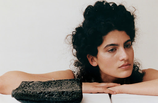 Portrait of a woman with curly dark hair resting her head on her hands, next to a porous black stone object against a white backdrop.