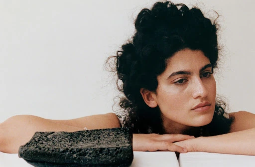 Portrait of a woman with curly dark hair resting her head on her hands, next to a porous black stone object against a white backdrop.