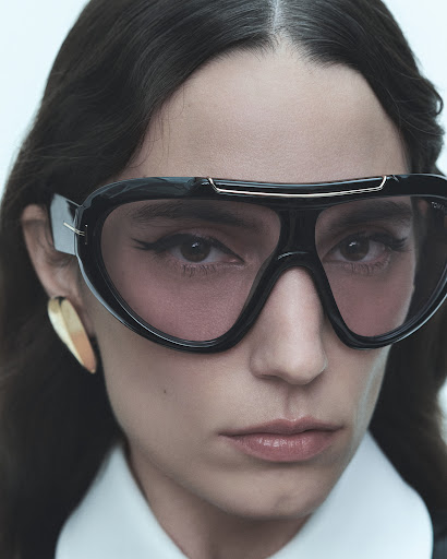 Close-up of a woman wearing oversized black sunglasses with pink lenses, gold earrings, and a white collared shirt against a white backdrop.