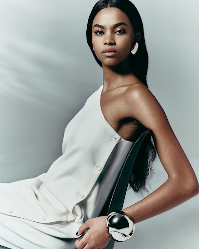 Stunning portrait of a woman with dark hair in a white asymmetrical dress and silver bangle, seated in a studio with soft lighting.