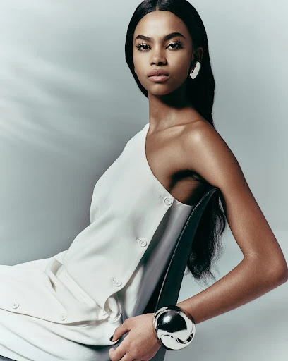 Stunning portrait of a woman with dark hair in a white asymmetrical dress and silver bangle, seated in a studio with soft lighting.