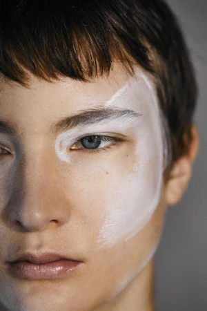 Close-up portrait of a model with short dark hair and abstract white face makeup painted around the eye.