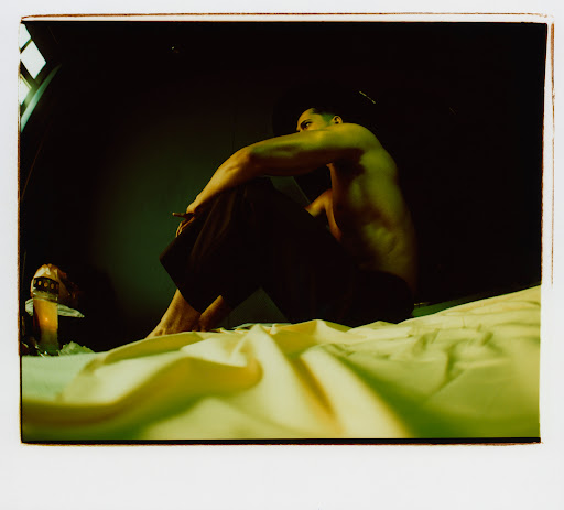 Bare-chested man sits on a bed holding a cigarette, with a decorative mushroom lamp visible; dark indoor setting.