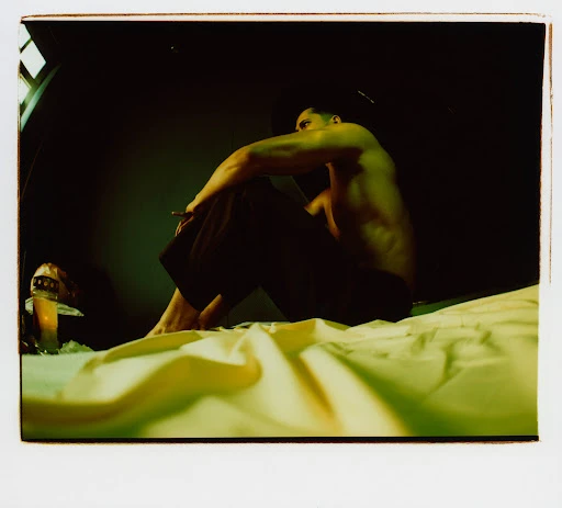 Bare-chested man sits on a bed holding a cigarette, with a decorative mushroom lamp visible; dark indoor setting.