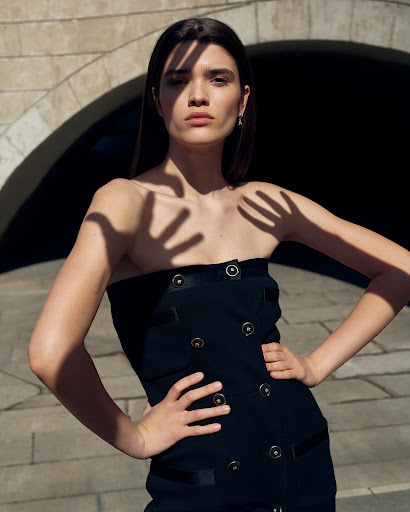 Portrait of a woman in a black dress with hands casting a shadow on her face, standing against a stone bridge background.