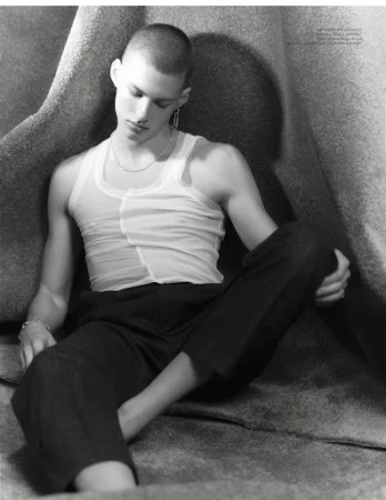 Monochrome portrait of a young man with a shaved head wearing a white tank top and dark trousers seated against a textured backdrop