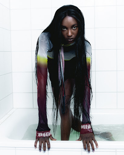 Dramatic portrait of a woman with long dark hair emerging from a bathtub filled with water, wearing a sheer printed top.